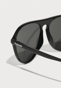 Ochelari de soare negri cu lentile întunecate, având un cadru din plastic elegant. Logo-ul Polaroid apare metalic pe latura bradului.