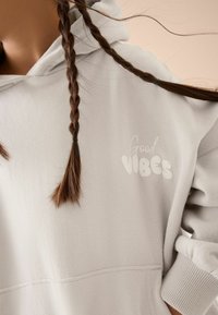 Ljusgrå hoodie med en mjuk textur, prydd med "Good Vibes" tryckt i en bubblig stil på bröstet. Inkluderar en huva med dragsko.