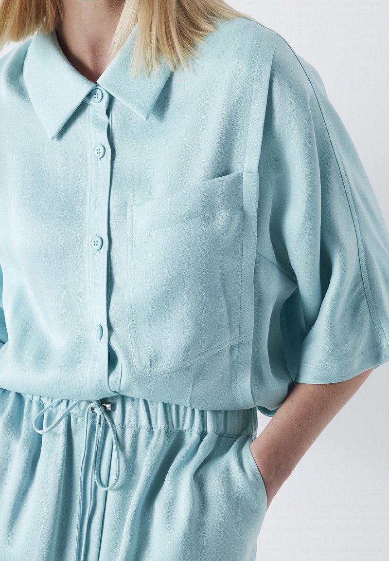 Blusa de manga corta azul claro con cuello, que cuenta con botones y un bolsillo en el pecho. Textura suave y lisa con cintura fruncida.