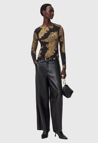 Pika mustega pikakäistega top, millel on kuldsed barokk-mustrid, koos laia jala mustade nahkpükstega. Mudel hoiab väikest musta kotti.