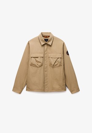 Beige Freizeitjacke mit langen Ärmeln und spitzem Kragen, zwei vorderen aufgesetzten Taschen mit Knopfklappe und rundem schwarzem Aufnäher am rechten Ärmel.