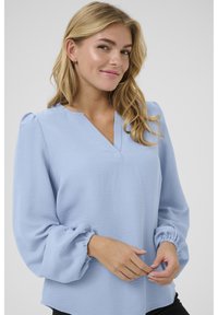 Blouse bleu clair avec un col en V, des manches bouffantes avec des poignets froncés, et un tissu texturé ; ourlet légèrement courbé sur les côtés.