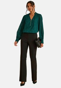 Blusa a maniche lunghe di colore teal con un motivo a onda testurizzato, colletto con zip, abbinata a pantaloni neri aderenti e una clutch nera. Calzature: tacchi neri.