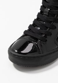 Svart lacklädersneaker med rund tå, texturerade svarta snören och en texturerad svart gummisåle. Glansig yta med sömdetalj.