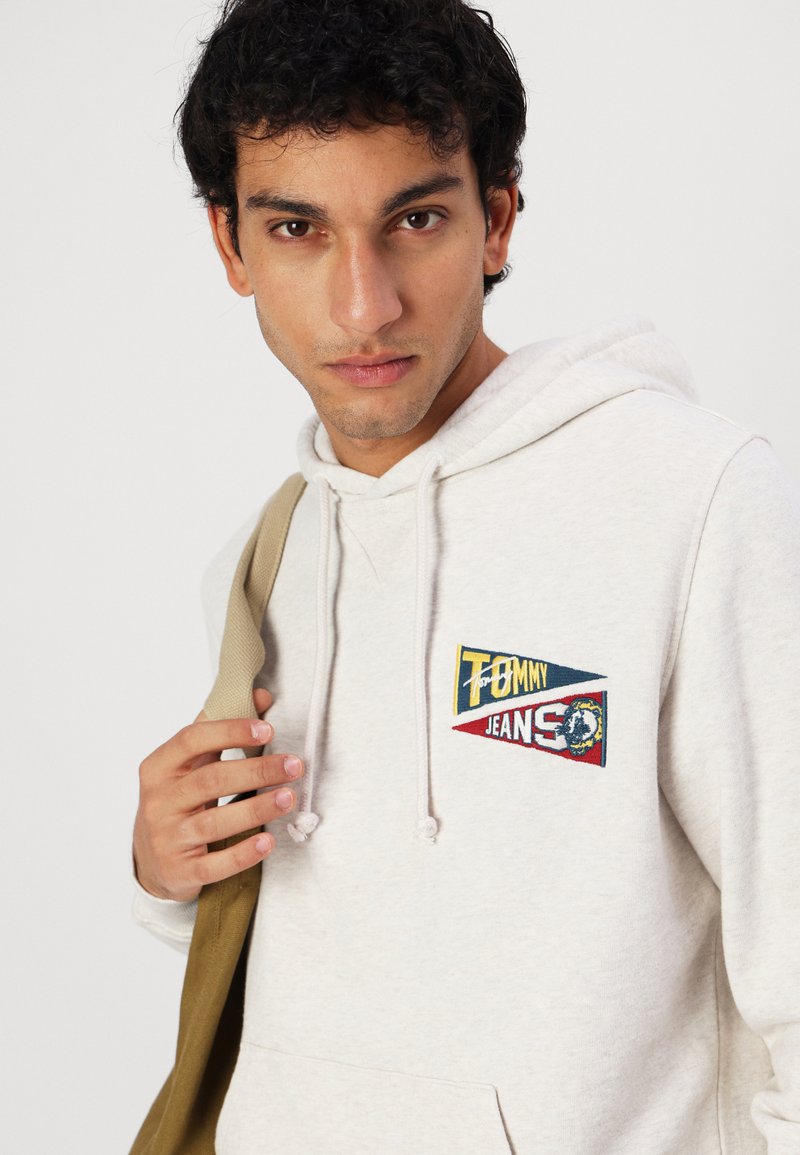 Lichtgrijze hoodie met een kangoeroezak, voorzien van geborduurde logo patches in blauw, rood en geel. Zachte katoenen stof met trekkoorden.