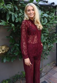 Bordó csipkeruha virágmintával, hosszú ujjakkal, galléros nyakkivágással és bő szabású nadrággal, zöld lombokkal a háttérben.