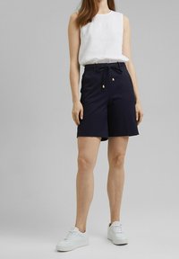 Pantalones cortos azul marino con cintura ajustable, detalles dorados y un corte relajado, combinados con una camiseta sin mangas blanca y zapatillas blancas.