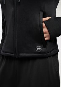 Giacca sportiva nera con zip frontale, maniche textured e applicazione del logo vicino all'orlo. Presenta tasche e una finitura in tessuto liscia.