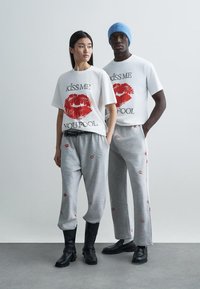 Bílé trička s červeným otiskem rtěnky a textem, doplněná šedými tepláky s rozptýlenými motivy rtěnky. Černé boty dotvářejí vzhled.