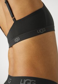 Svart bh med glatt stoff, justerbare stropper og bredt bånd med UGG-merke i grått. Designet for en tettsittende passform.