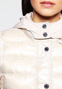 Světlý béžový prošívaný kabát s vysokým límcem a knoflíkovým zapínáním. Hladká, lesklá látka s horizontálním prošíváním a odnímatelnou kapucí.