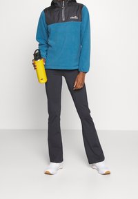 Pullover in pile blu con dettagli neri, colletto con zip e logo. Il modello indossa pantaloni scuri e tiene in mano una bottiglia d'acqua gialla. Sneakers bianche.