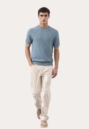 Man loopt naar voren en draagt een lichtblauwe textuur korte-mouwtrui, beige broek en bruine instapschoenen tegen een neutrale achtergrond.