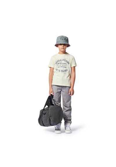 Grijze bucket hat, crèmekleurig graphic t-shirt, lichtgrijze broek en witte sneakers. Houdt een donkergrijze duffeltas met een gestructureerd oppervlak vast.