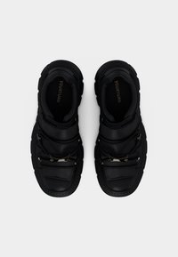 Ζευγάρι από μαύρα chunky αθλητικά παπούτσια με παχιά σόλα, μεταλλικές λεπτομέρειες με αγκράφες και λουράκια βέλκρο, που φαίνονται από ψηλά σε λευκό φόντο.