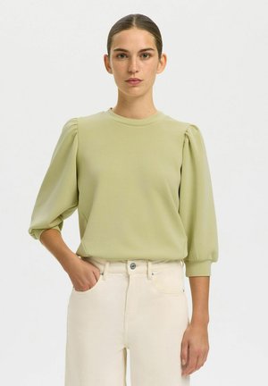 Kvinne med brunt hår samlet bak, iført en lysegrønn bluse med puffede ermer og kremfargede bukser med høy midje, står med den ene hånden i lommen.