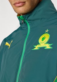 En turkise vindjakke med grøn lynlås, prydet med et gult Puma-logo og et broderet klubbadge i grøn og gul med en stjerne og bånd detaljer.