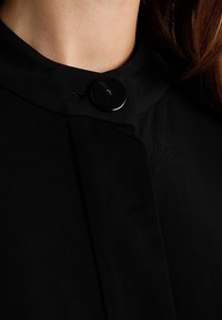 Indumento con colletto nero caratterizzato da una texture liscia, con un bottone nero rotondo e un design elegante che enfatizza la semplicità.