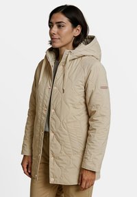 Beige gewatteerde jas met een capuchon, voorzien van roze drukknoopjes, een klepzak en gestructureerde golfpatronen op de stof.