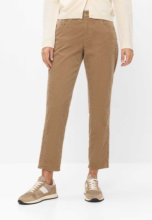 Nedre del af en person iført lysebrune manchesterbukser, beige og hvide sneakers og en cremefarvet sweater mod en hvid baggrund.