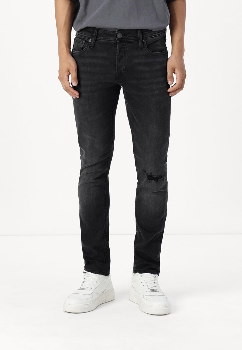 Jeans de mezclilla negros con un corte ajustado, que presentan un detalle desgastado en la rodilla y un estilo clásico de cinco bolsillos. Combinados con zapatillas blancas.