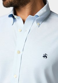 Světle modrá košile na tlačítka vyrobená z látky s jemnou texturou. Obsahuje límec, uzávěr na knoflíky a malé vyšívané logo v námořnické modři.