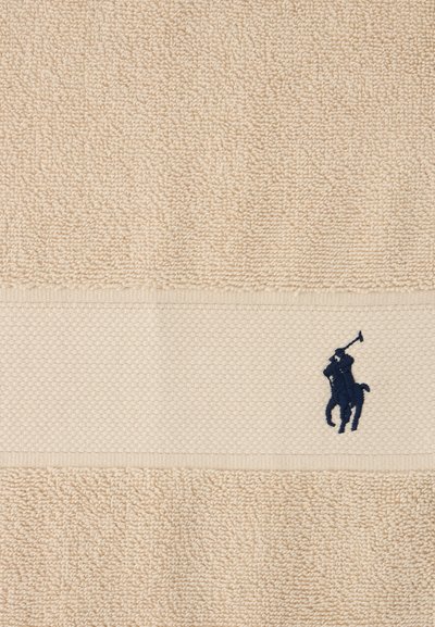 Μπεζ πετσέτα με υφή και μια οριζόντια ταινία που δείχνει έναν μαύρα κεντημένο παίκτη πόλο πάνω σε άλογο που κρατά ένα υψωμένο ρόπαλο.