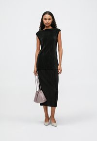 Γυναίκα με μακριά σγουρά μαλλιά, φορώντας ένα μαύρο αμάνικο τοπ και αντίστοιχη φούστα midi, κρατώντας μία μικρή μωβ τσάντα και φορώντας λευκά μυτερά παπούτσια.