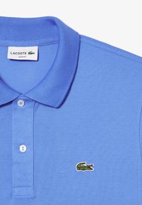 Slim fit blå pikétröja, tillverkad av texturerad tyg. Har ett birdeye-mönster, tre-knapps knäppning och en liten grön broderad krokodil-logotyp.