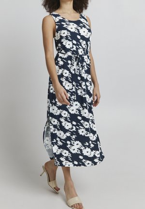 Kobieta ubrana w bezrękawnikową, granatową sukienkę midi z białym kwiatowym wzorem, z bocznymi rozcięciami, w beżowych butach na obcasie z odkrytymi palcami, stojąca na jednolitym tle.