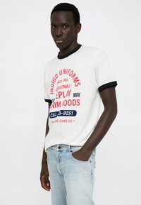 Λευκό μπλουζάκι με κόκκινο και μπλε κείμενο, μαύρο γιακά και κοντά μανίκια. Ανοιχτό μπλε τζιν με σκισίματα και μια τσέπη μπροστά.