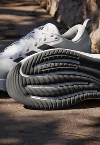 Chaussure de sport blanche avec logo noir, lacets et semelle noire texturée reposant sur le sol extérieur près des racines d'un arbre.