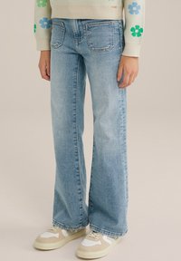 Ljusblå utsvängda jeans med femficksdesign, kontrasterande sömmar och en mjuk denimtextur; bärs med beige och vita sneakers.