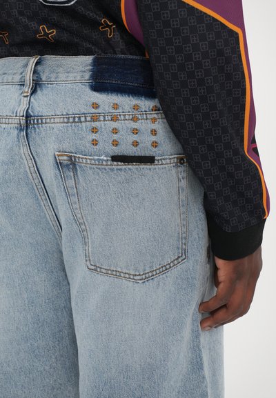 Jeans din denim de culoare albastru deschis, cu un finisaj uzat, având cusături decorative marron și modele geometrice pe buzunarul din spate.
