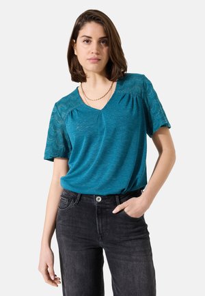 Top corto de color azul verde con detalles de encaje en los hombros, escote en V y ajuste relajado, combinado con jeans negros, que muestran una textura suave.