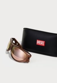 Óculos de sol castanhos com uma armação texturizada e lentes espelhadas cor-de-rosa, acompanhados de uma caixa preta da marca Diesel.