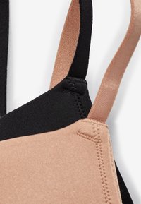 Gros plan d'une bretelle de soutien-gorge beige et noire montrant la texture du tissu et les détails des coutures sur fond blanc.