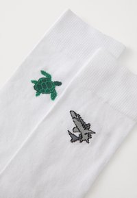 Chaussettes blanches avec un design de tortue verte sur une chaussette et un design de requin gris sur l'autre, fabriquées en tissu doux et texturé.