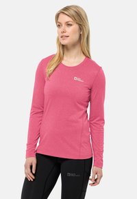 Frau in einem pinkfarbenen Langarmshirt von Jack Wolfskin und schwarzen Leggings von Jack Wolfskin, die mit neutralem Gesichtsausdruck vor einem weißen Hintergrund steht.