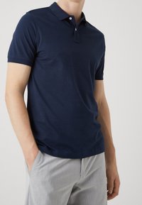 Mann trägt ein navyblaues kurzärmliges Poloshirt und hellgraue Hosen, mit einer Hand in seiner Tasche, vor einem einfarbigen Hintergrund.