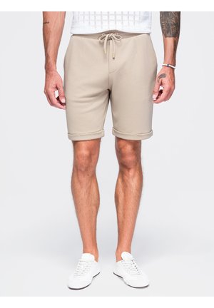 Mand iført beige shorts med snor og hvide sneakers, stående med armene afslappet ned langs siden på en ensartet lys baggrund.