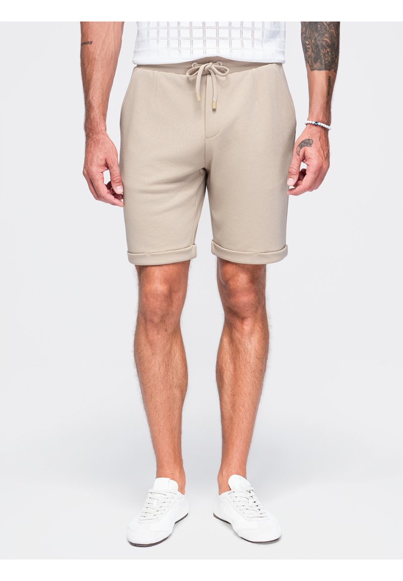 Homme portant un short beige à cordon et des baskets blanches, debout avec les bras détendus le long du corps sur un fond clair uni.