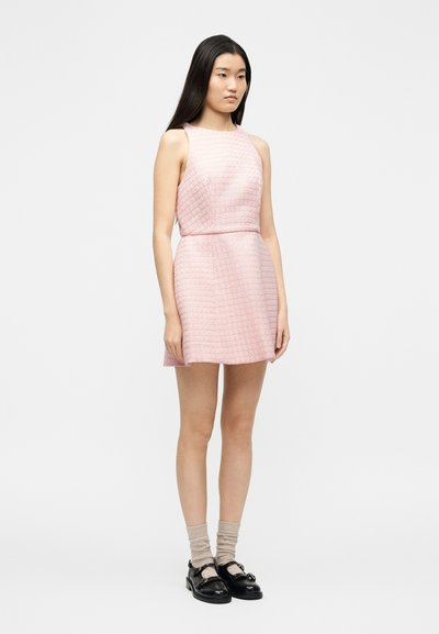 Νεαρή γυναίκα με μακριά μαύρα μαλλιά, φορώντας ένα ροζ αμάνικο μίνι φόρεμα με υφή, μπεζ κάλτσες και μαύρα παπούτσια με αγκράφα, στέκεται πλάγια.