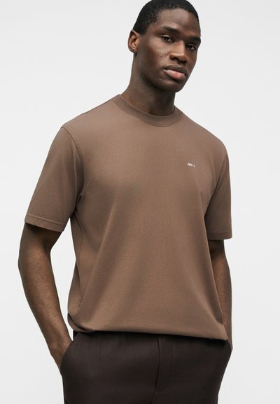 Νεαρός άνδρας με σκουρόχρωμο δέρμα που φορά καφέ κοντομάνικο πουκάμισο και σκούρο παντελόνι, στέκεται με τα χέρια στις τσέπες μπροστά σε λευκό φόντο.