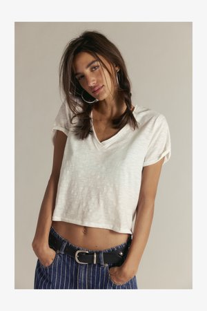 Jauna moteris su tamsiais plaukais surištais dviem kasomis, dėvinti didelius žiedinius auskarus, baltą trumpą V formos kaklo marškinėlį ir mėlynas dryžuotas džinsines kelnes, rankas kišenėse.