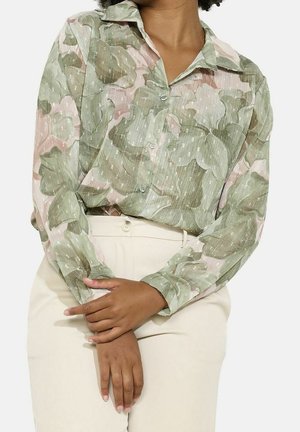 Persona con una camisa abotonada con estampado floral verde y rosa y pantalones crema de tiro alto, con las manos suavemente entrelazadas al frente.