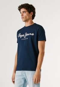 Junger Mann mit lockigem Haar, der ein marineblaues "Pepe Jeans London" T-Shirt und hellblaue Jeans trägt und vor einem schlichten hellen Hintergrund steht.