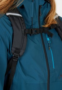 Blågrön vattentät jacka med svarta ryggsäcksremmar. Har en framdragkedja med turkosa detaljer och en reflekterande axelaccent.