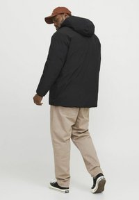 Zwarte hoodie met een gladde textuur, gecombineerd met beige broek en zwarte sneakers. De pet is bruin, wat een contrasterend detail toevoegt.