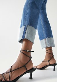 Vaqueros de mezclilla con puño azul claro, dobladillo sin acabar y corte recto, combinados con sandalias negras de tira con tacón cuadrado y correa en el tobillo.